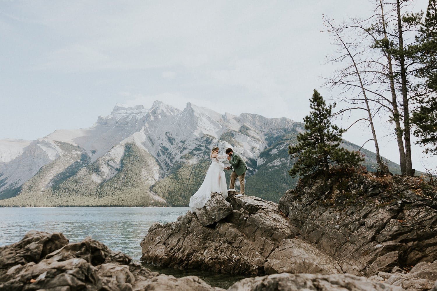 groom kisses brides hand on the shoreline at lake minnewanka in Banff Alberta